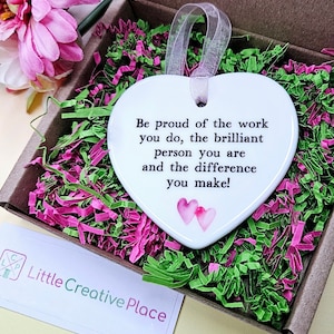 May include: A white heart-shaped ornament with a ribbon, inside a cardboard box filled with pink and green shredded paper. The ornament has the text "Be proud of the work you do..." and two pink hearts. A small card is also visible.