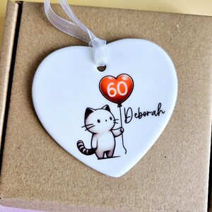 May include: A white heart-shaped ornament with a cartoon cat holding a red balloon with the number 60. The name "Deborah" is written in cursive. A white ribbon is attached for hanging.