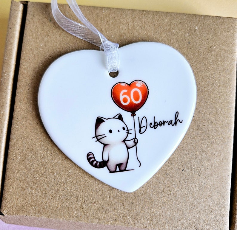 May include: A white heart-shaped ornament with a cartoon cat holding a red balloon with the number 60. The name "Deborah" is written in cursive. A white ribbon is attached for hanging.