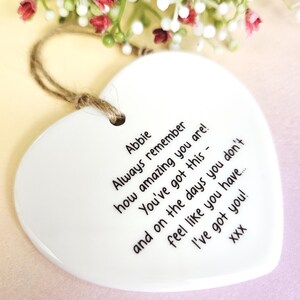 May include: A white heart-shaped ceramic ornament with a jute string for hanging. The ornament has black text that reads "Abbie, Always remember how amazing you are! You've got this - and on the days you don't feel like you have... I've got you!" Decorative flowers are in the background.