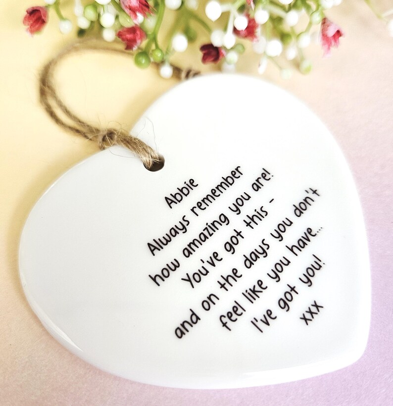 May include: A white heart-shaped ceramic ornament with a jute string for hanging. The ornament has black text that reads "Abbie, Always remember how amazing you are! You've got this - and on the days you don't feel like you have... I've got you!" Decorative flowers are in the background.