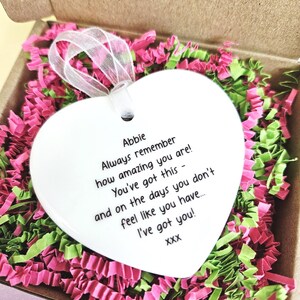 May include: A white heart-shaped ornament with a ribbon, inside a cardboard box filled with pink and green shredded paper. The ornament has the text "Abbie Always remember how amazing you are! You've got this - and on the days you don't feel like you have... I've got you!"