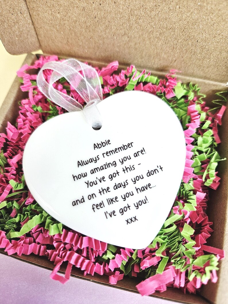 May include: A white heart-shaped ornament with a ribbon, inside a cardboard box filled with pink and green shredded paper. The ornament has the text "Abbie Always remember how amazing you are! You've got this - and on the days you don't feel like you have... I've got you!"
