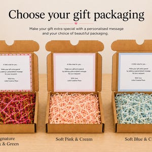 Three open cardboard gift boxes, each filled with shredded paper in different colours and a personalised message card. The boxes are labelled "Signature Pink & Green", "Soft Pink & Cream", and "Soft Blue & Cream". Text at the top reads "Choose your gift packaging".