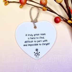 May include: White heart-shaped ceramic ornament with a jute string for hanging. The ornament features the text "A truly great team is hard to find, difficult to part with and impossible to forget!" and a small floral design.
