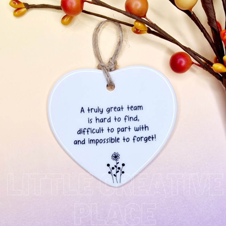 May include: White heart-shaped ceramic ornament with a jute string for hanging. The ornament features the text "A truly great team is hard to find, difficult to part with and impossible to forget!" and a small floral design.
