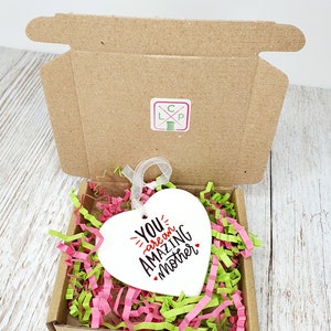 May include: White ceramic heart-shaped ornament with the text "You are an amazing mother" in black lettering with a red heart accent. The ornament is in a small cardboard box with pink and green shredded paper.