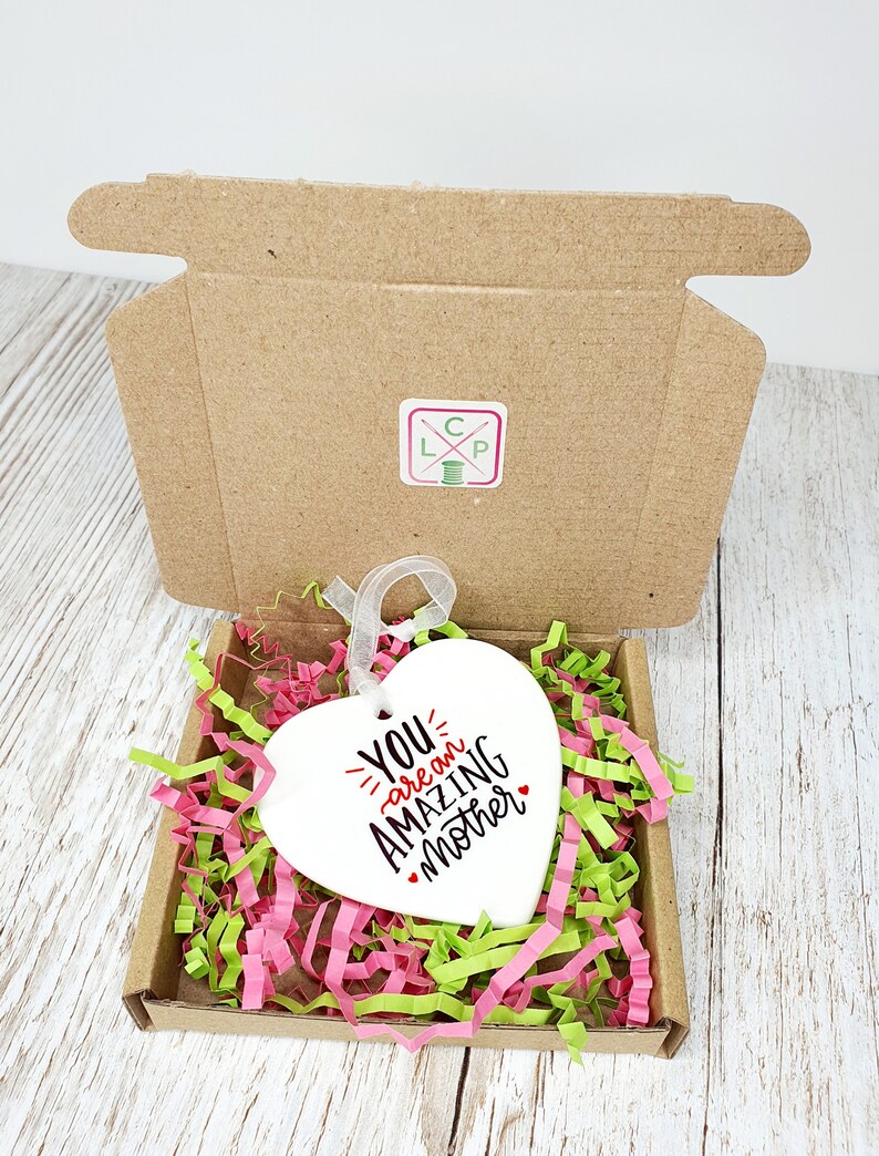May include: White ceramic heart-shaped ornament with the text "You are an amazing mother" in black lettering with a red heart accent. The ornament is in a small cardboard box with pink and green shredded paper.