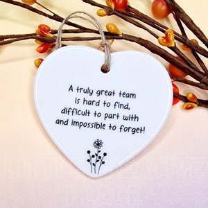 May include: White heart-shaped ceramic plaque with a jute string for hanging. The plaque features the text "A truly great team is hard to find, difficult to part with and impossible to forget!" and a small floral design.