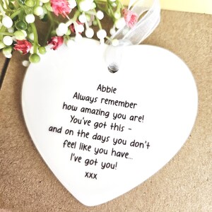 May include: A heart-shaped ceramic ornament with a ribbon, featuring the name "Abbie" and an inspirational message in black text. The background includes small white and pink flowers, adding a delicate touch to the design.