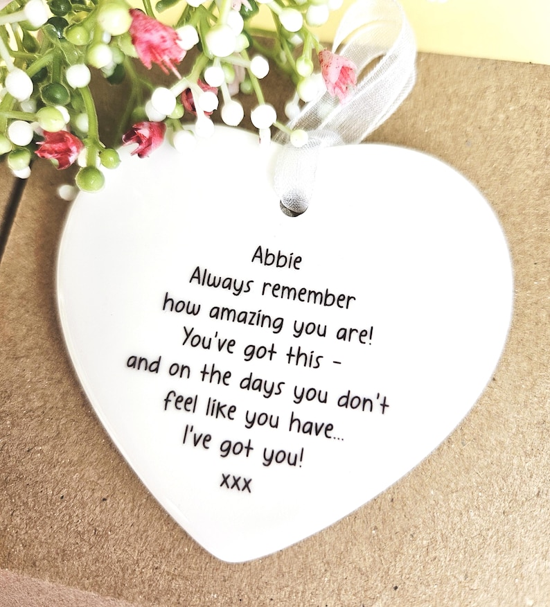 May include: A heart-shaped ceramic ornament with a ribbon, featuring the name "Abbie" and an inspirational message in black text. The background includes small white and pink flowers, adding a delicate touch to the design.
