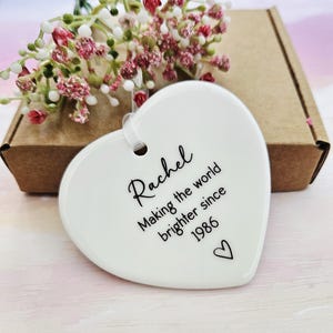 White ceramic heart keepsake personalised with name and special year, handmade birthday ornament displayed with kraft gift box and soft floral background.