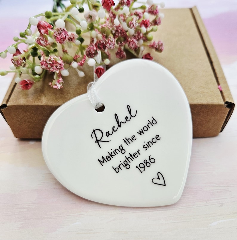 White ceramic heart keepsake personalised with name and special year, handmade birthday ornament displayed with kraft gift box and soft floral background.