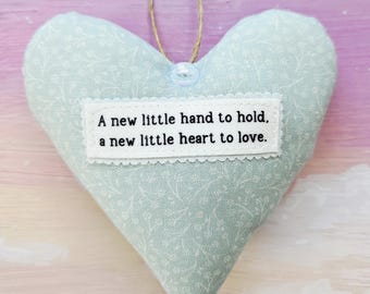May include: A light blue heart-shaped fabric ornament with a floral pattern. The ornament has a white rectangular patch with the text "A new little hand to hold, a new little heart to love." A twine loop is attached for hanging.