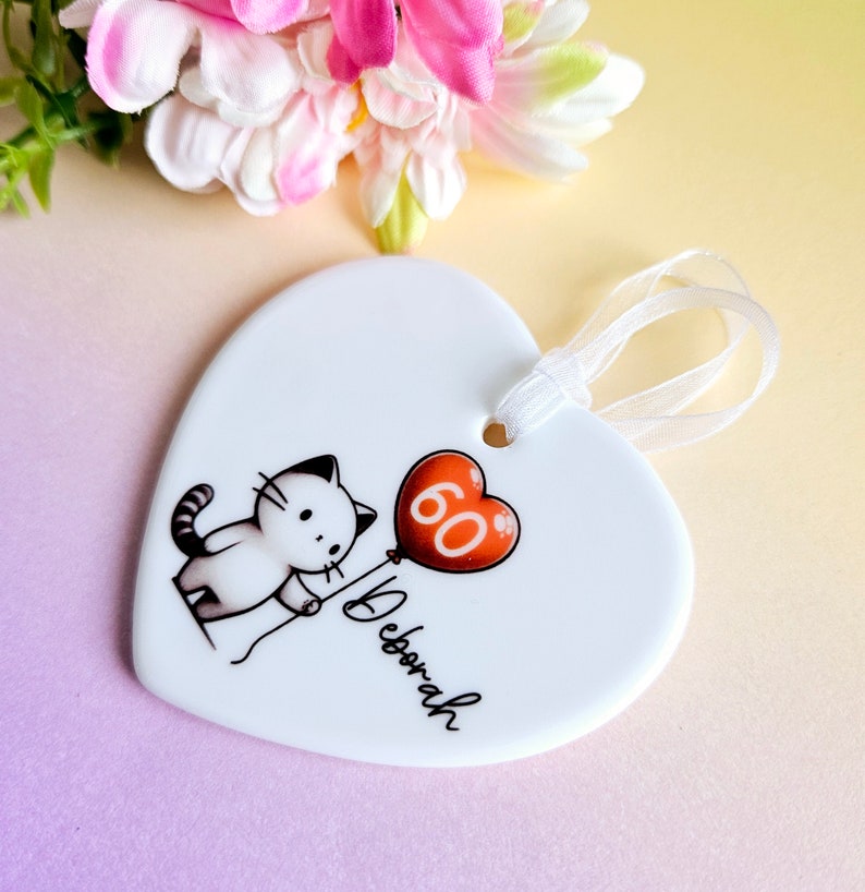 May include: White heart-shaped ornament with a cartoon cat holding a red balloon with the number 60. The name "Deborah" is written below the cat. A white ribbon is attached for hanging.
