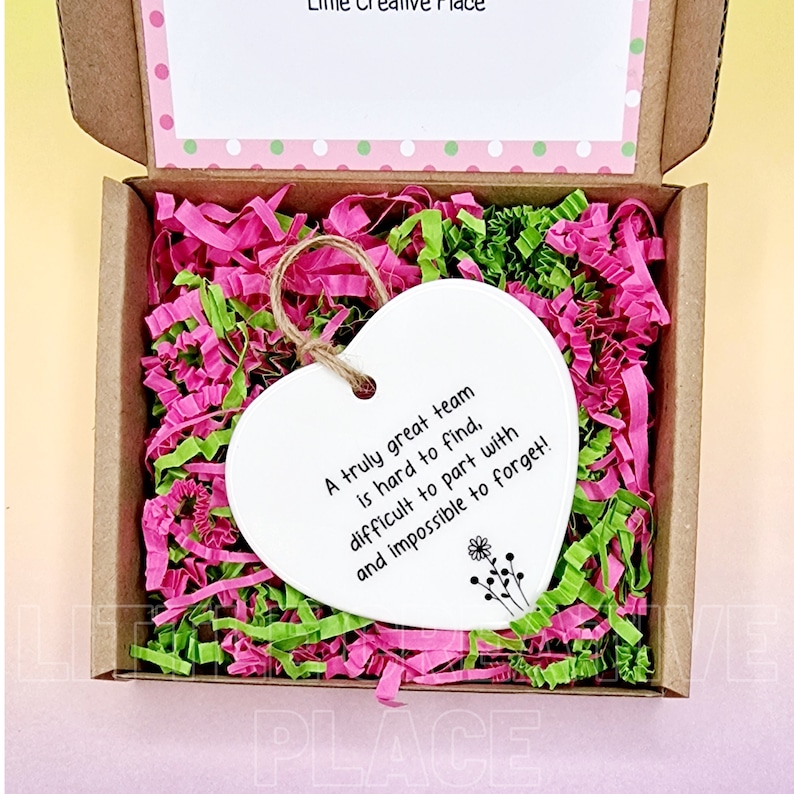 May include: A heart-shaped white ornament with a jute string, inside a cardboard box filled with pink and green shredded paper. The ornament has the text "A truly great team is hard to find, difficult to part with and impossible to forget!"