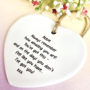 May include: A white heart-shaped ceramic ornament with a jute string for hanging. The ornament has black text that reads "Always remember how amazing you are! You've got this - and on the days you don't feel like you have... I've got you!" and the name "Abbie".