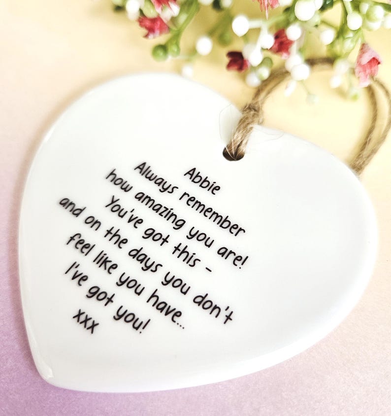 May include: A white heart-shaped ceramic ornament with a jute string for hanging. The ornament has black text that reads "Always remember how amazing you are! You've got this - and on the days you don't feel like you have... I've got you!" and the name "Abbie".