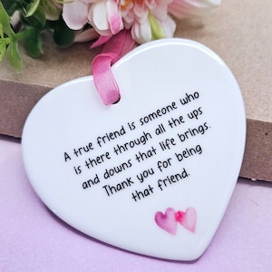 May include: White ceramic heart-shaped ornament with a pink ribbon. The ornament has a message that reads: "A true friend is someone who is there through all the ups and downs that life brings. Thank you for being that friend."