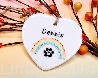 May include: White heart-shaped ceramic ornament with the name "Dennis" in black. A colorful rainbow and a paw print design are below the name. The ornament has a jute string for hanging. Decorative branches with orange berries are in the background.