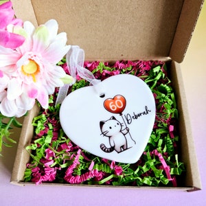 May include: A white heart-shaped ornament with a cartoon cat holding a red balloon with the number 60. The name "Deborah" is written in cursive. The ornament is in a cardboard box with pink and green paper filler, and a pink flower.