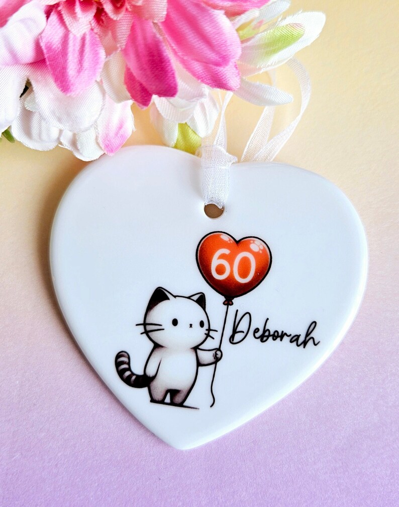 May include: A white heart-shaped ornament with a cartoon cat holding a red balloon with the number 60. The name "Deborah" is written in script. A white ribbon is attached for hanging. Pink and white flowers are in the background.