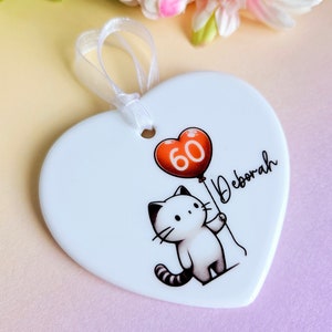 May include: White heart-shaped ornament with a cartoon cat holding a red balloon with the number 60. The name "Deborah" is written in script. A white ribbon is attached for hanging.