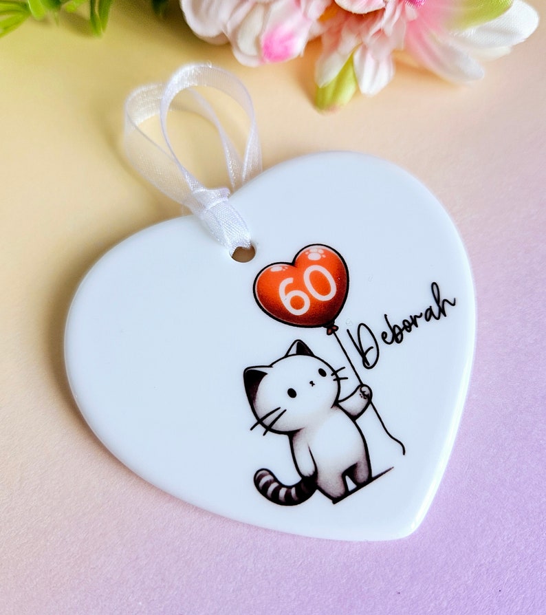 May include: White heart-shaped ornament with a cartoon cat holding a red balloon with the number 60. The name "Deborah" is written in script. A white ribbon is attached for hanging.
