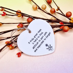 May include: A white heart-shaped ceramic ornament with a jute string for hanging. The ornament has black text that reads, "A truly great team is hard to find, difficult to part with and impossible to forget!" Decorative floral accents are also present.