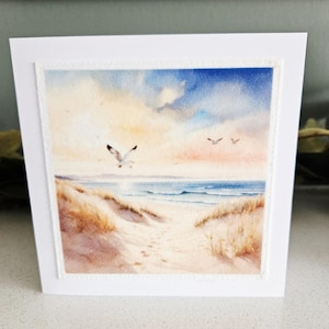 May include: A white greeting card with a watercolour painting of a beach scene. The artwork shows a sandy path leading to the sea, with seagulls flying in a blue and cloudy sky. The card has a white border.