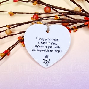 May include: A white heart-shaped ceramic ornament with a jute string for hanging. The ornament has the text "A truly great team is hard to find, difficult to part with and impossible to forget!" and a small floral design.