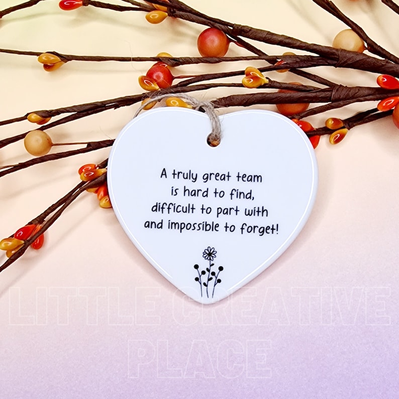 May include: A white heart-shaped ceramic ornament with a jute string for hanging. The ornament has the text "A truly great team is hard to find, difficult to part with and impossible to forget!" and a small floral design.