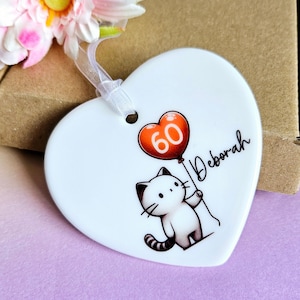 May include: White heart-shaped ornament with a cartoon cat holding a balloon with the number 60. The name "Deborah" is written in cursive. A white ribbon is attached for hanging. The background is a mix of purple and brown.