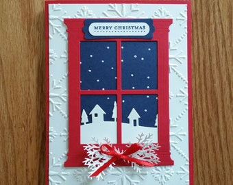 May include: A handmade Christmas card featuring a red window frame with a snowy scene. The window reveals a dark blue sky with falling snow and white silhouettes of houses and trees. The card has a white embossed snowflake background and a red bow.