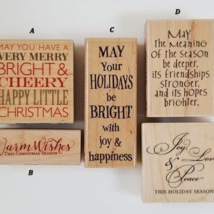 May include: A collection of wooden stamps with holiday-themed text in various fonts. The stamps feature phrases like "Merry Christmas," "Warm Wishes," and "Joy, Love & Peace." The text is in red and black.