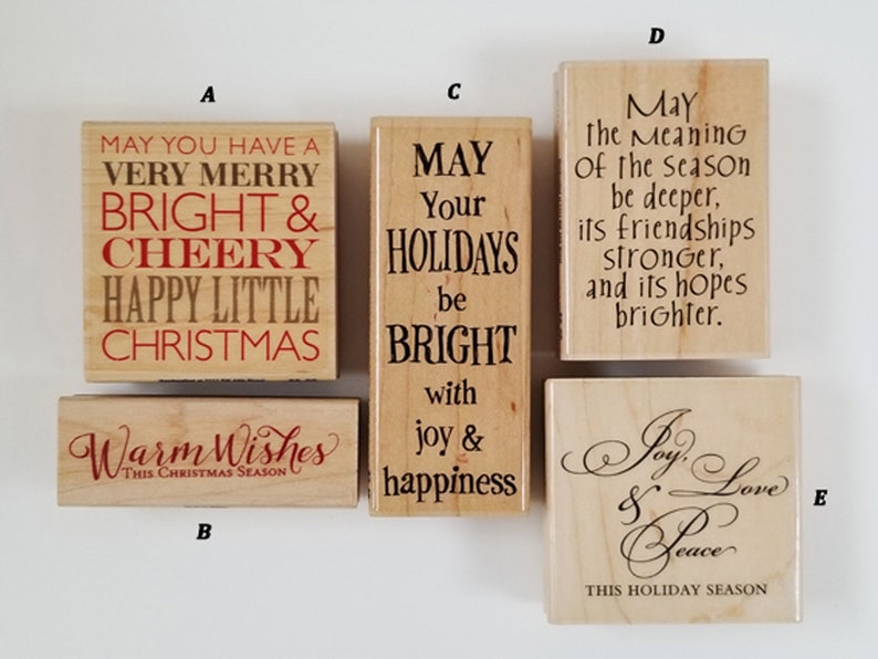 May include: A collection of wooden stamps with holiday-themed text in various fonts. The stamps feature phrases like "Merry Christmas," "Warm Wishes," and "Joy, Love & Peace." The text is in red and black.