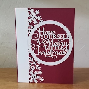 Puede incluir: Una tarjeta de Navidad con fondo burdeos y una franja vertical blanca a la izquierda. La tarjeta presenta un diseño circular blanco con las palabras "Have Yourself A Merry Little Christmas" recortadas. Copos de nieve blancos decoran el lado izquierdo.