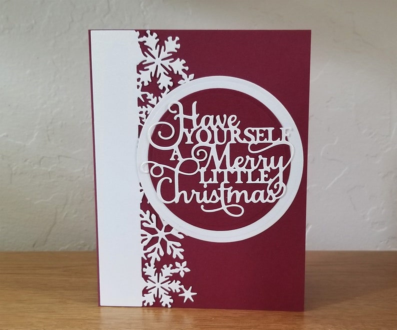 May include: A Christmas card with a burgundy background and a white vertical stripe on the left. The card features a white circular design with the words "Have Yourself A Merry Little Christmas" cut out. White snowflakes decorate the left side.