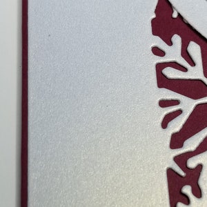 May include: Close-up of a white, textured paper with a decorative cut-out design in a deep red color. The design has a floral or snowflake-like pattern. The edge of the paper is a deep red.