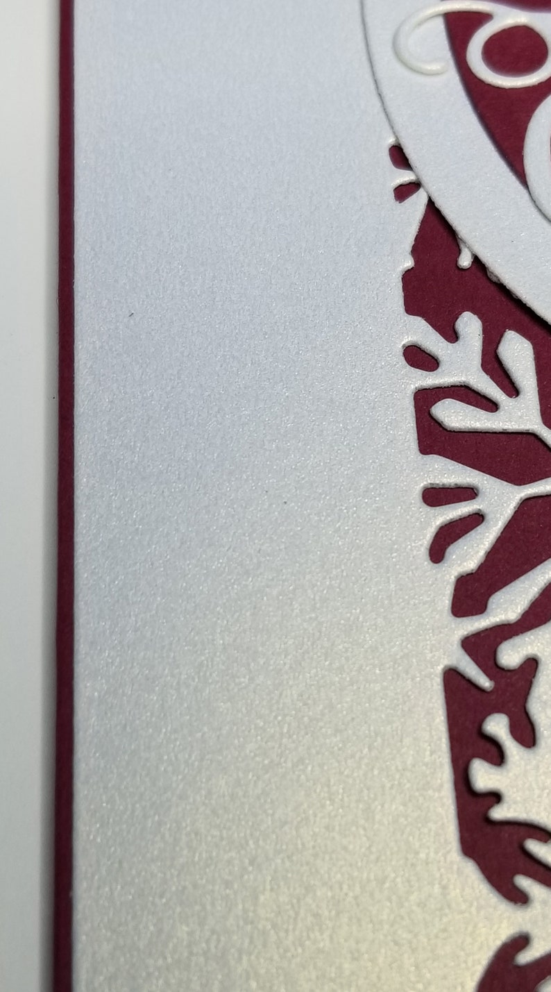May include: Close-up of a white, textured paper with a decorative cut-out design in a deep red color. The design has a floral or snowflake-like pattern. The edge of the paper is a deep red.