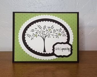 May include: A green card with a black border and a white circle with a black outline. Inside the circle is a black tree with green leaves and three small green birds. The text "With Sympathy" is written in black on a white label with a black outline.