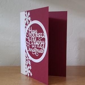 May include: A folded Christmas card with a burgundy cover and a white interior. The front features a white oval with the words "Have Yourself a Merry Little Christmas" cut out. White snowflakes decorate the left side.