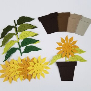 以下が含まれることがあります： ひまわり、葉、植木鉢の切り絵。ひまわりは黄色とオレンジ色の中心部、葉は緑色、植木鉢は茶色です。切り絵は白い背景に配置されています。