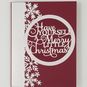 May include: A Christmas card with a burgundy background and a white border on the left side. The card features a circular cutout with the words "Have Yourself A Merry Little Christmas" and snowflake cutouts.
