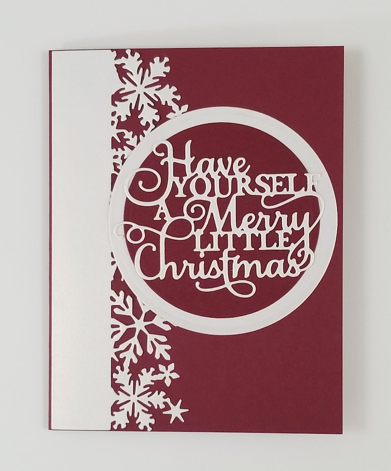 May include: A Christmas card with a burgundy background and a white border on the left side. The card features a circular cutout with the words "Have Yourself A Merry Little Christmas" and snowflake cutouts.