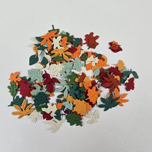 Puede incluir: Un conjunto de hojas de papel recortadas de colores, de diversas formas y tamaños. Las hojas son en tonos naranja, rojo, verde y crema, lo que sugiere un tema otoñal. Ideal para proyectos de manualidades o scrapbooking.