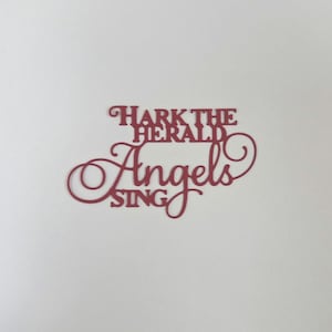 May include: A burgundy-colored cutout with the words "Hark the Herald Angels Sing" in a decorative font. The text is arranged in three lines, with the word "Angels" in a flowing script. The cutout is set against a white background.