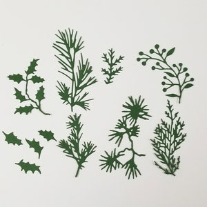 May include: A selection of dark green paper cutouts depicting botanical elements such as holly leaves, pine needles, and berry branches. The cutouts are arranged on a white background, ideal for crafting or festive decorations.