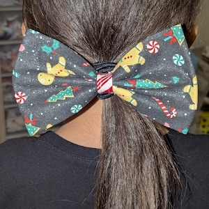 May include: A dark gray fabric bow with a Christmas-themed pattern, including gingerbread men, Christmas trees, and candy canes. The bow is secured with a black elastic band with a red and white striped accent.