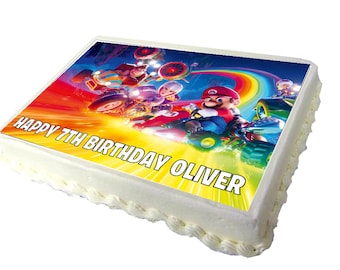 May include: A white cake with a colorful Mario Kart themed edible image on top. The image features Mario, Donkey Kong, and Peach racing on a rainbow track. The text "Happy 7th Birthday Oliver" is printed on the cake.